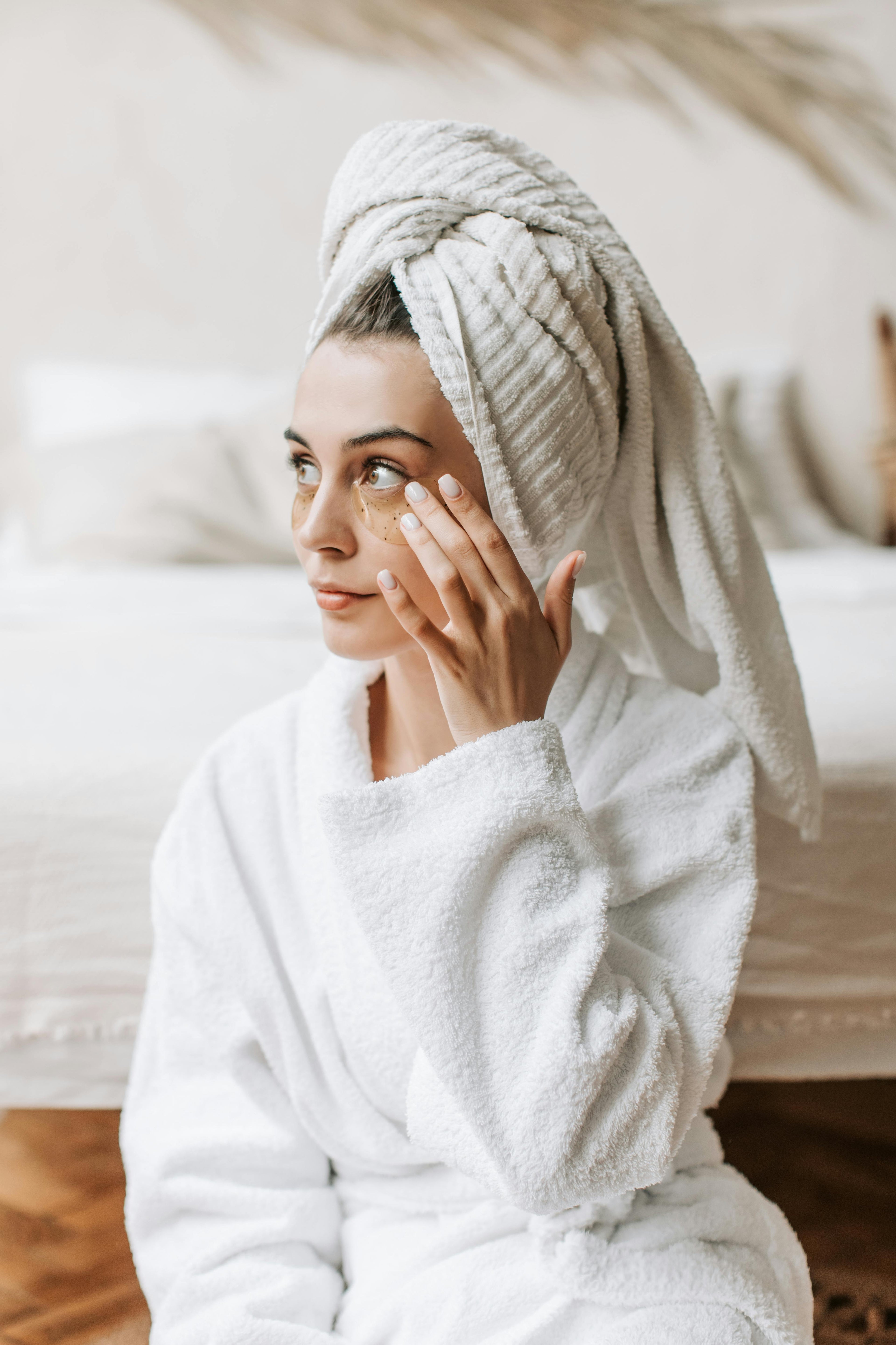 Woman in white bathrobe applying under-eye patches as part of her skincare routine. Self-care moment with The Koko Collection.