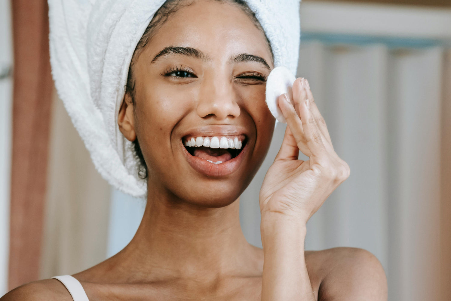 Smiling woman cleansing her face with a cotton pad and towel wrapped around her hair