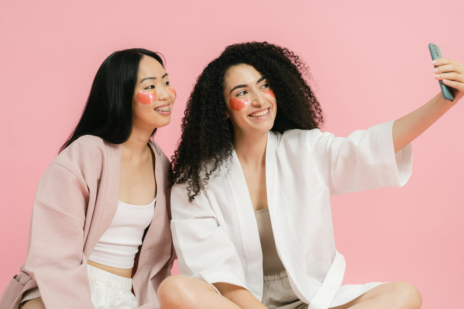 Skincare besties in robes with eye patches smiling for a selfie on a pink backdrop
