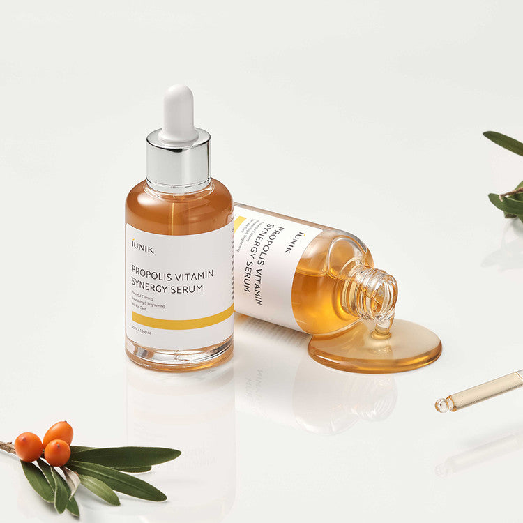 Two bottles of iUNIK Propolis Vitamin Synergy Serum with spilled product and sea buckthorn, showcasing nourishing and brightening Korean skincare.