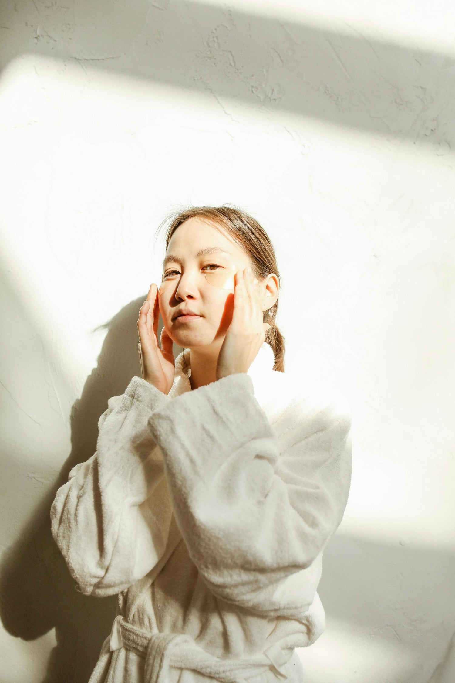 K-beauty morning skincare moment with woman in bathrobe applying product in soft sunlight