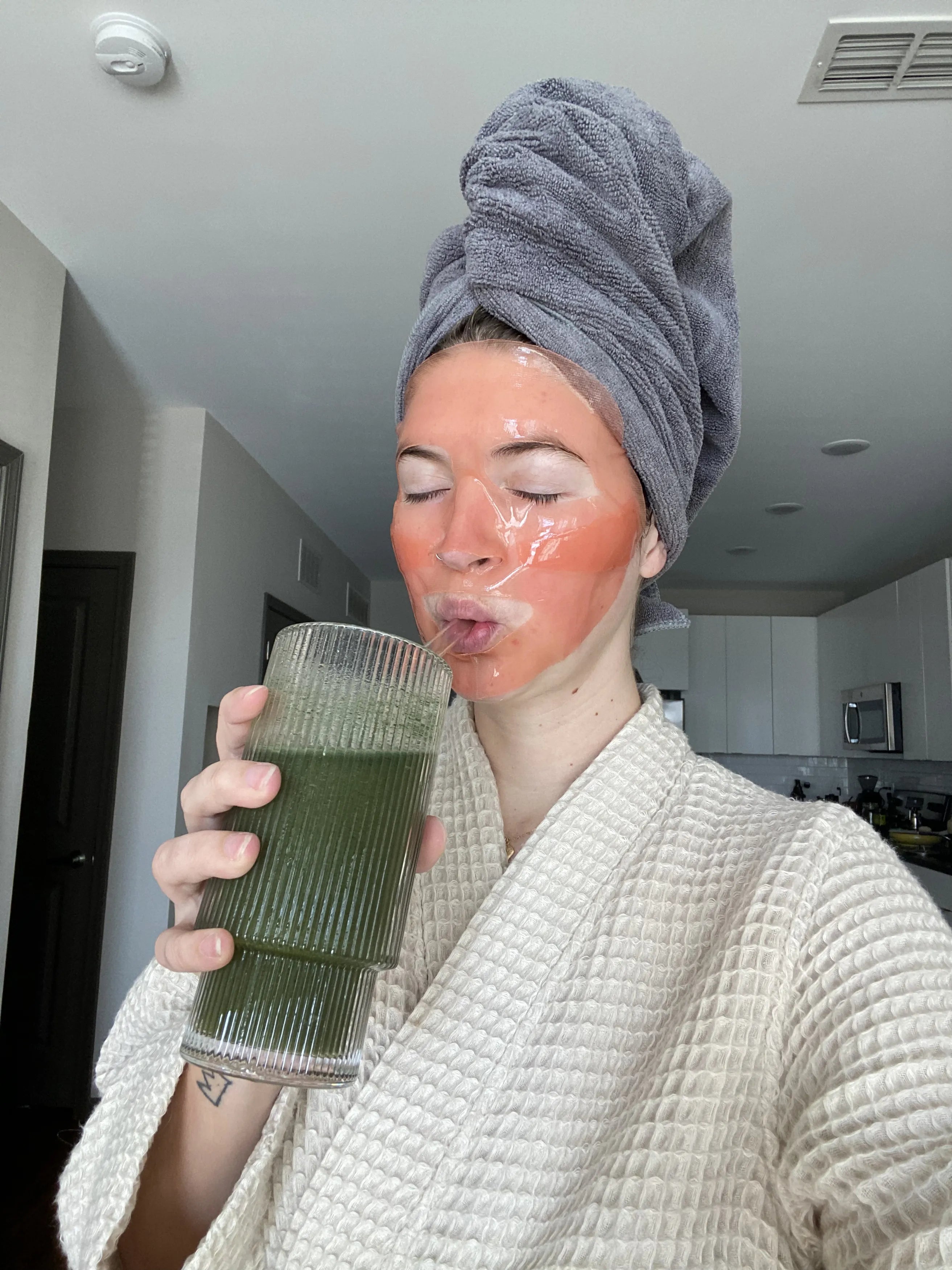 Woman in a robe with a towel wrapped around her head, wearing a hydrogel face mask and drinking a green smoothie—capturing a self-care skincare moment.