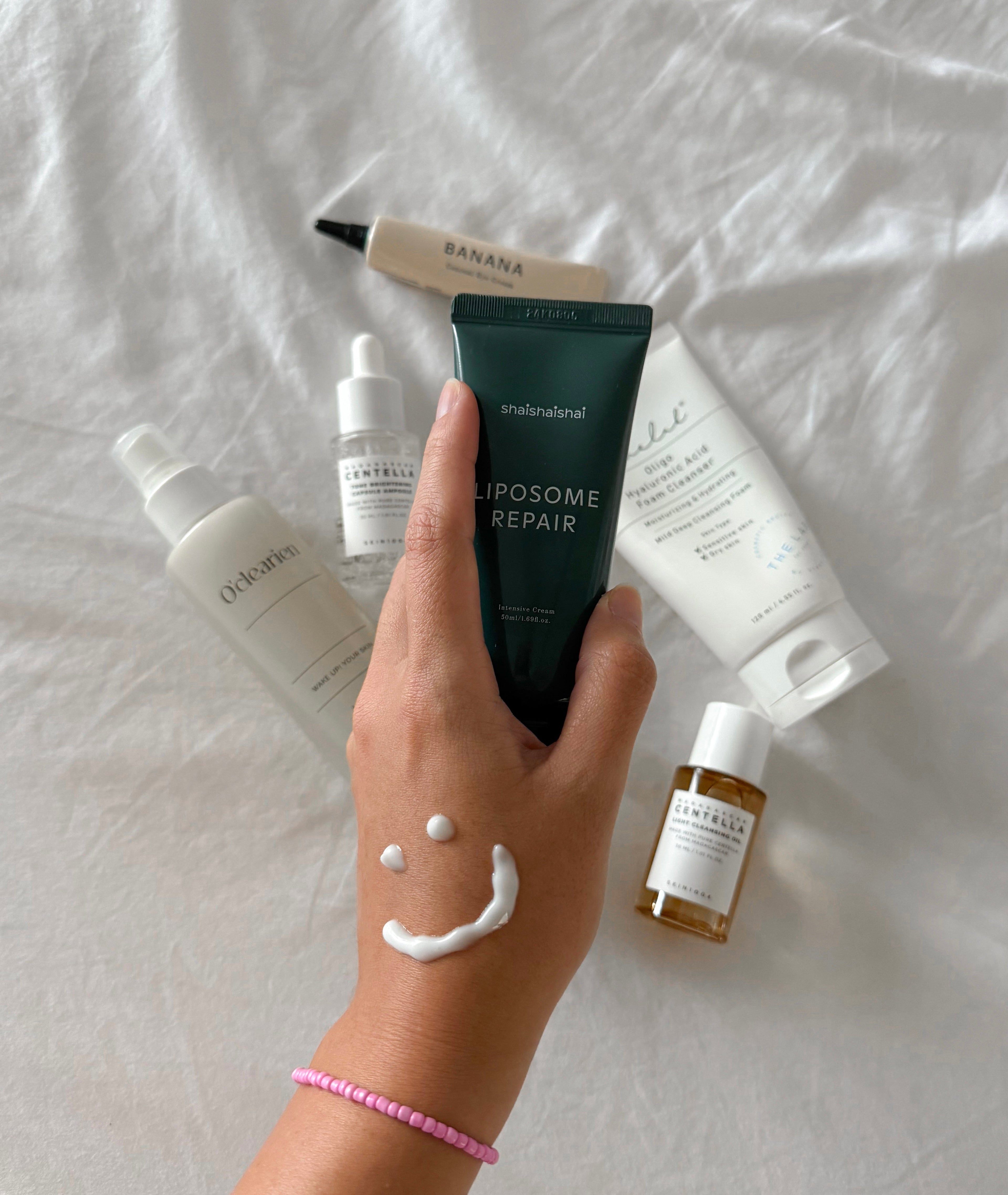 Playful skincare flat lay showing hand with smiley made of cream, holding Liposome Repair cream, surrounded by K-beauty serums, oils, and cleansers