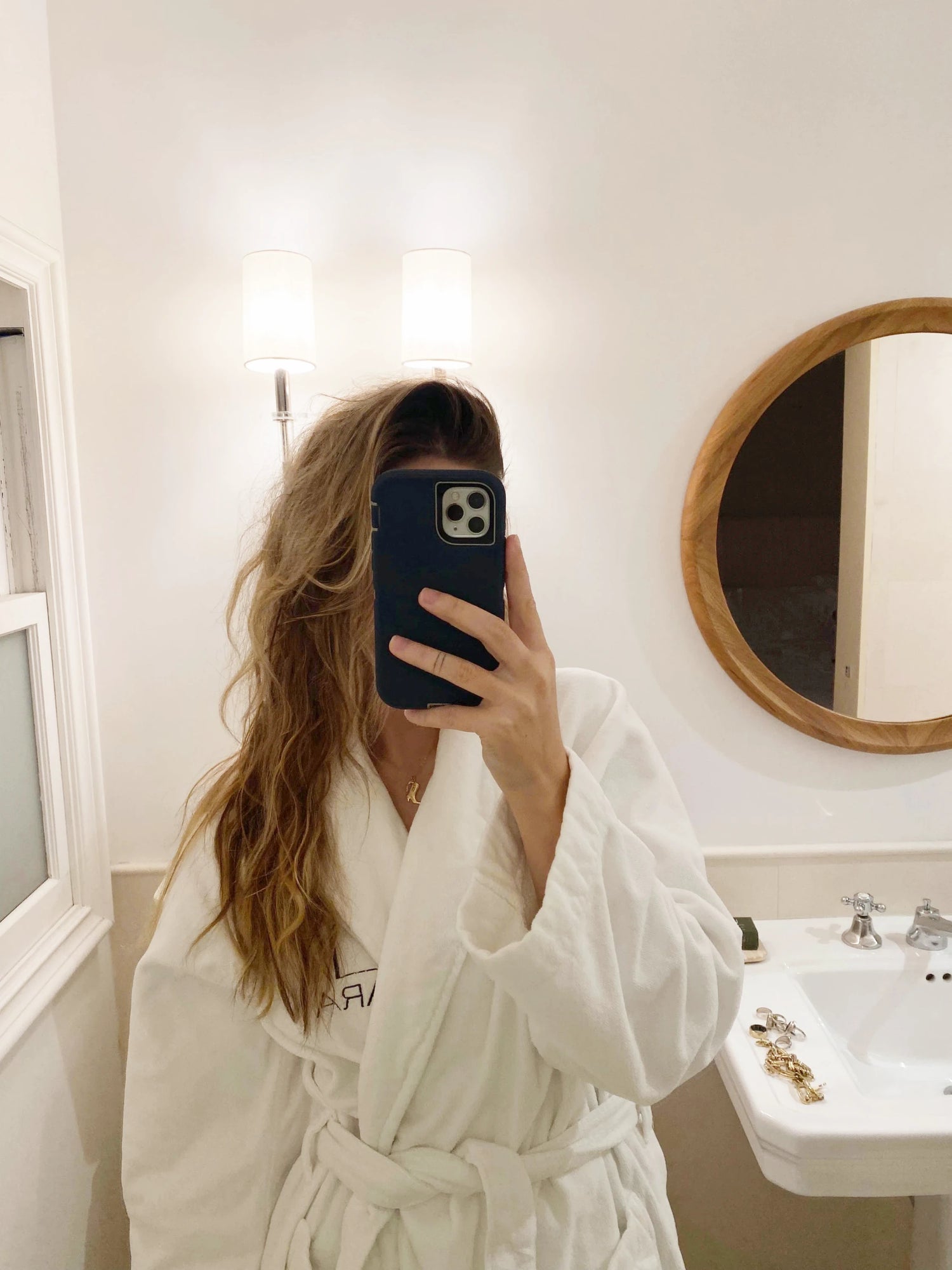 Mirror selfie of a woman in a white robe during her nighttime routine, standing in a softly lit bathroom with minimal decor, shared by The Koko Collection.