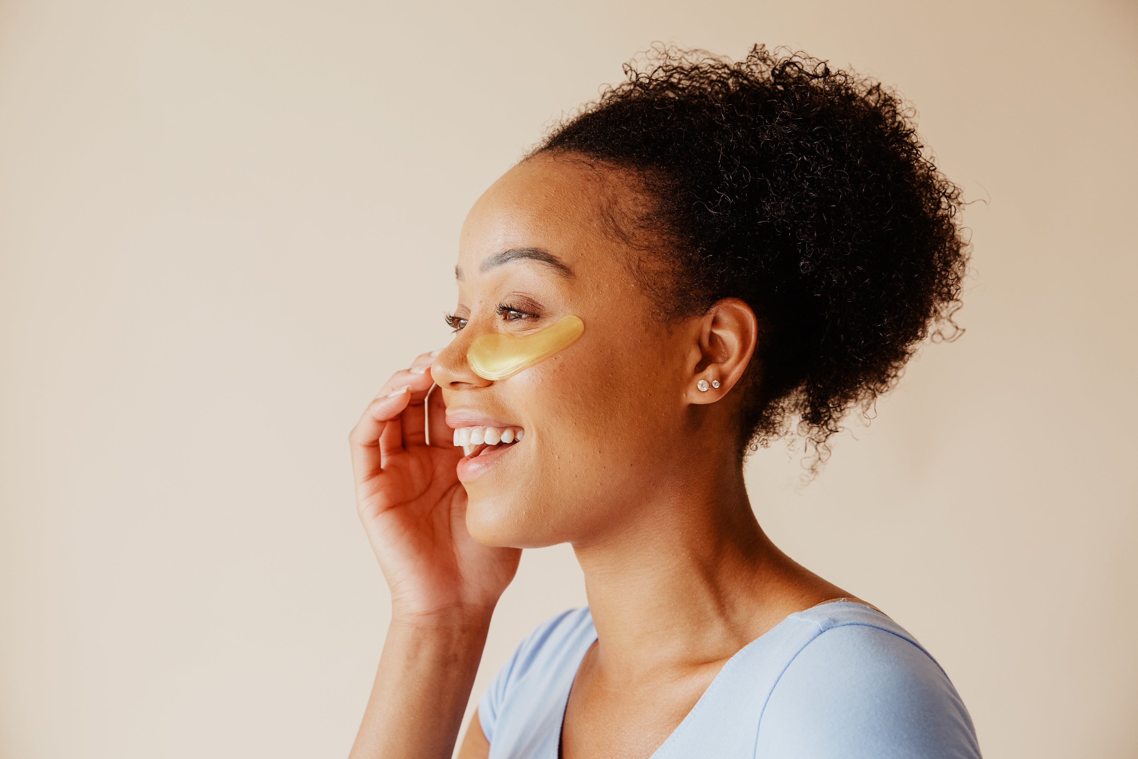 Smiling woman applying golden under-eye patches for skincare, promoting glow-boosting Korean beauty essentials from The Koko Collection.